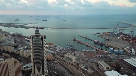 Aerial-view-of-downtown-Haifa-landmark-sail-tower,-sea-port-and-bay-water,-Israel,-overhead-circle-drone