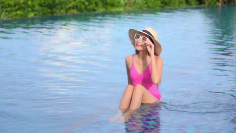 young attractive asian woman in pink swimsuit and white sunglasses sitting on edge of pool enjoying sunbathing