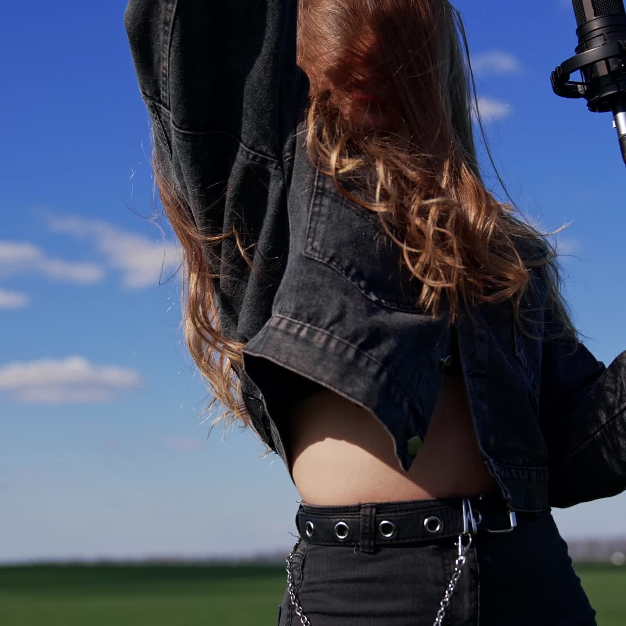 Cantante pop donna vicino al microfono all'aperto. Bella ragazza in tuta di jeans che scuote la testa con i capelli lunghi mentre canta su sfondo di campo verde.