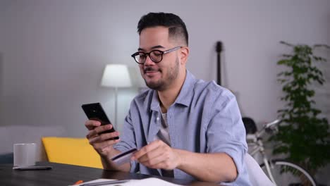 joven asiático usando teléfono inteligente con tarjeta de crédito para compras en línea y pagos en casa.
