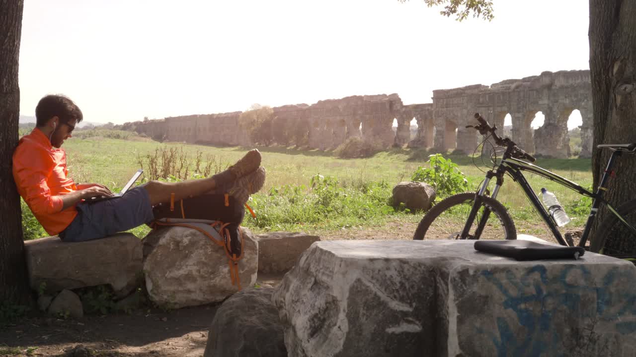 고대 로마의 수로 앞에서 나무 의 바위 위에 앉아 노트북으로 글을 쓰고 있는 자전거 타는 젊은 매력적인 운동선수, 오렌지색 스포츠 옷과 배을 입은 남자, 로마의 파르코 델리 아쿠에도티 (parco degli acquedotti)