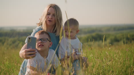 a woman in a blue dress sits on the grass, smiling as she takes a picture with her smartphone. her two sons, both wearing white shirts, are with her; while the older one sits close