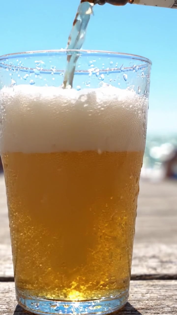 Rinfrescante bevanda dorata versata in un bicchiere, con bollicine che salgono e schiuma che si forma sotto un cielo azzurro brillante in una calda giornata di sole