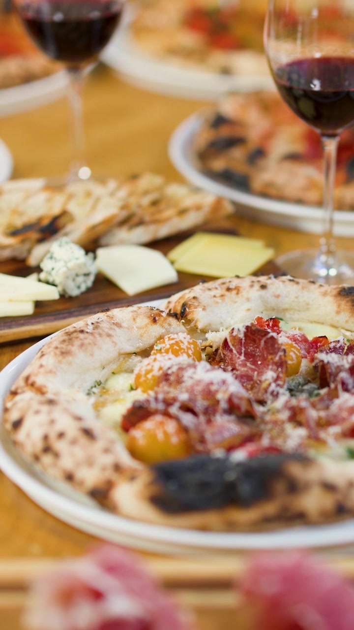 Un primo piano inclinato della telecamera rivela pizze artigianali, taglieri di salumi e vino rosso in un ristorante.