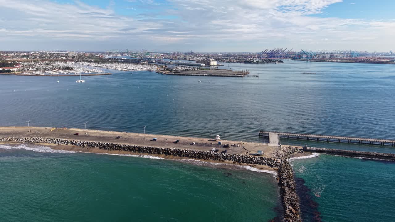 Aerial drone view of harbor entrance with breakwater, pier, and coastal city skyline in background