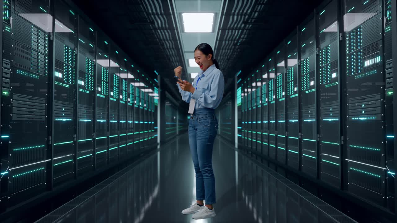 Vista laterale a figura intera di una donna asiatica con il suo smartphone in un data center, alza il pugno con un grido di vittoria.