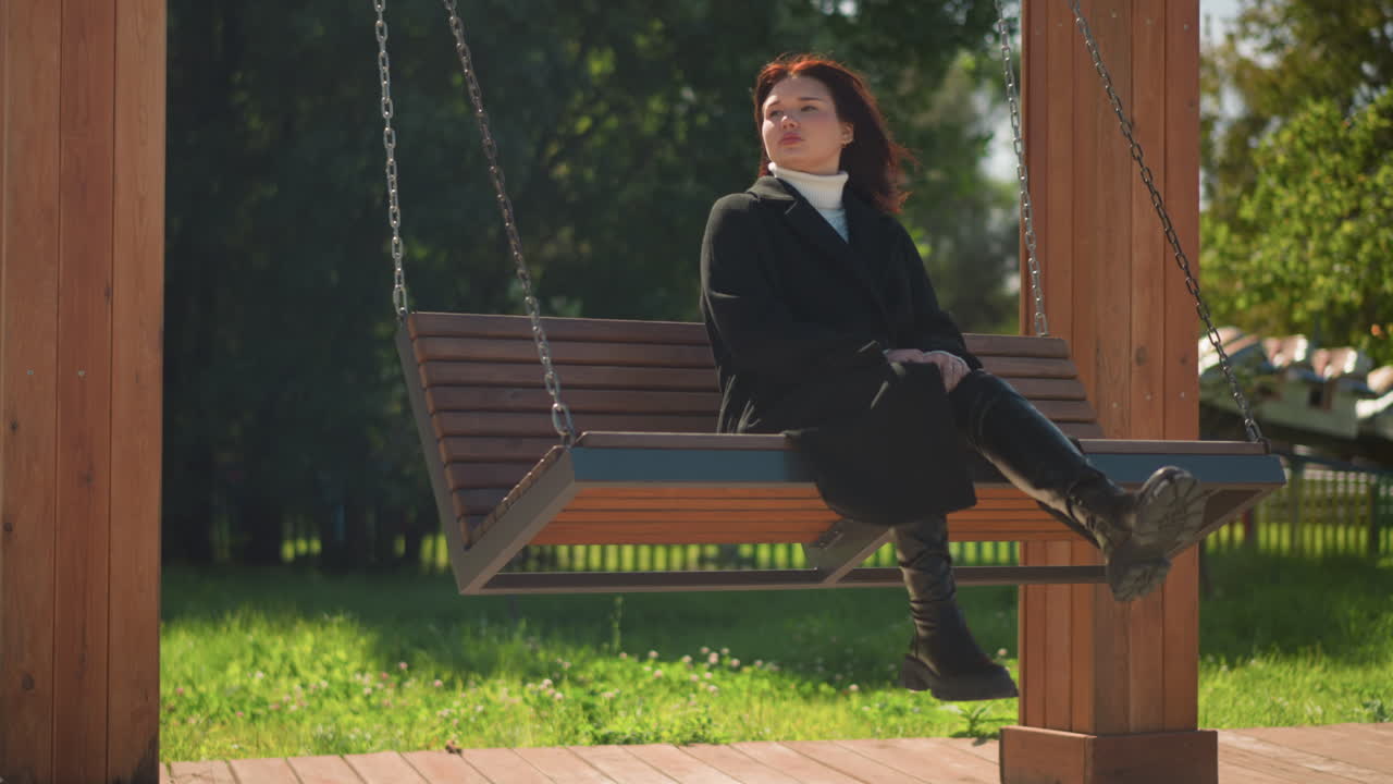 Stylish woman in black coat and boots serenely sits on wooden swing, hands clasped between legs, gazing into the distance in a sunlit par