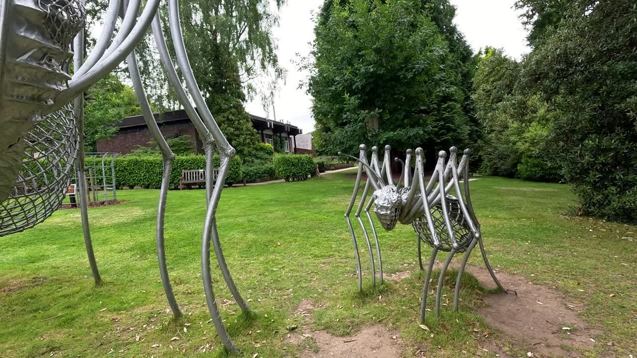 A handheld camera circles a large metallic spider sculpture outdoors in Durham’s botanical gardens, capturing intricate details and lush greenery under diffuse daylight