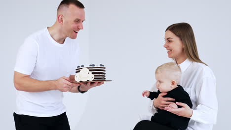 smiling mom in white shirt with blond baby in hands. happy man comes up with a cake approaching to a child. white backdrop.