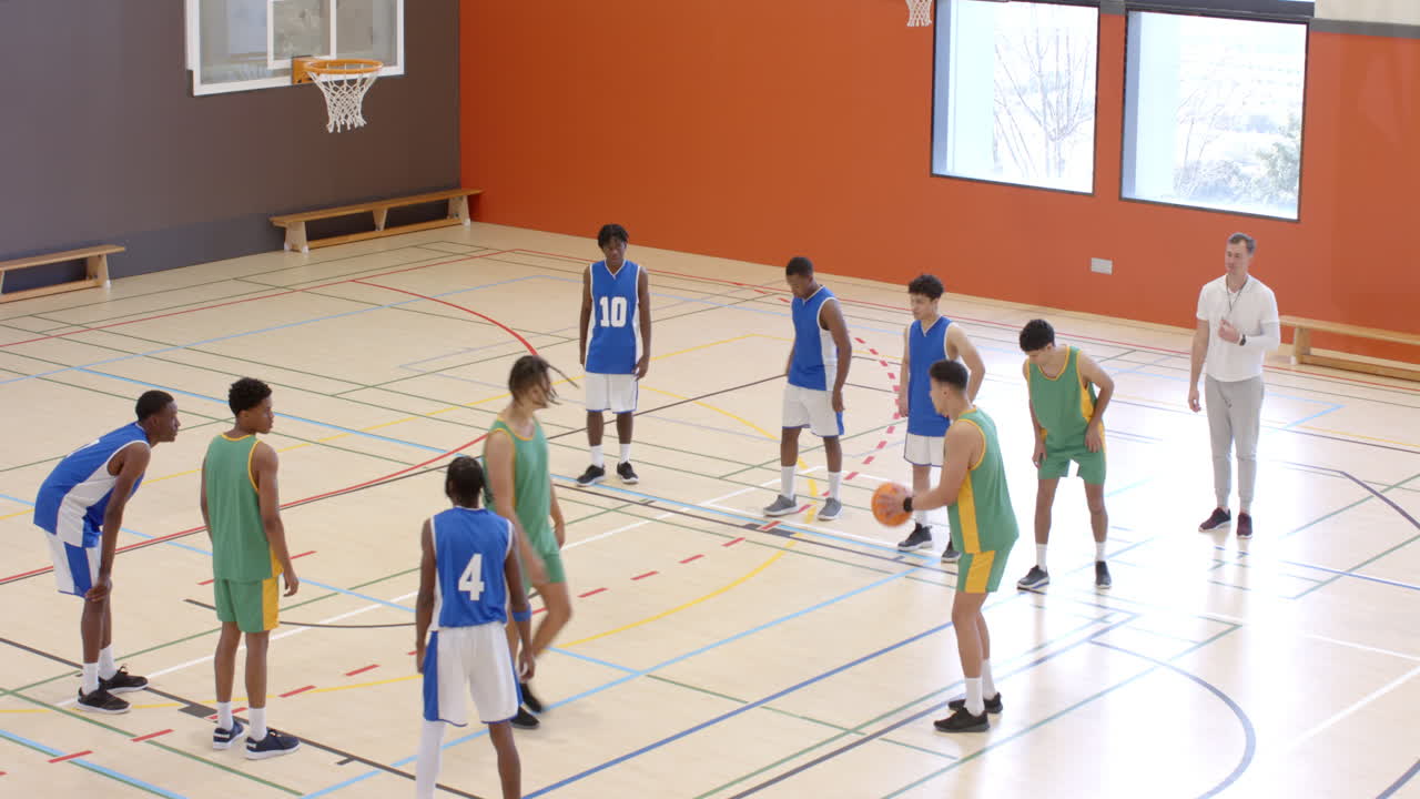 Multiracial male basketball players in gym and throwing ball into basket with coach observing game