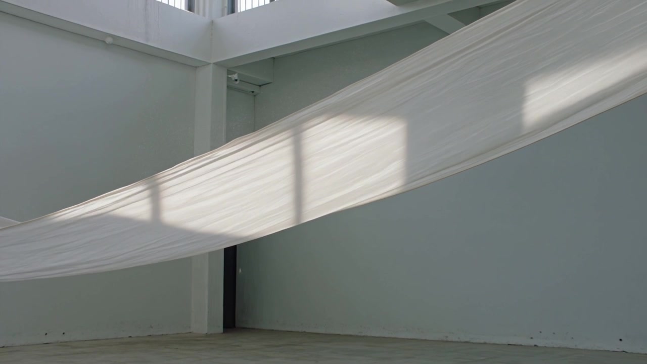Luce solare che si sposta dalle finestre a cleristorio, muovendosi su un drappo bianco trasparente in una galleria, causando leggere increspature