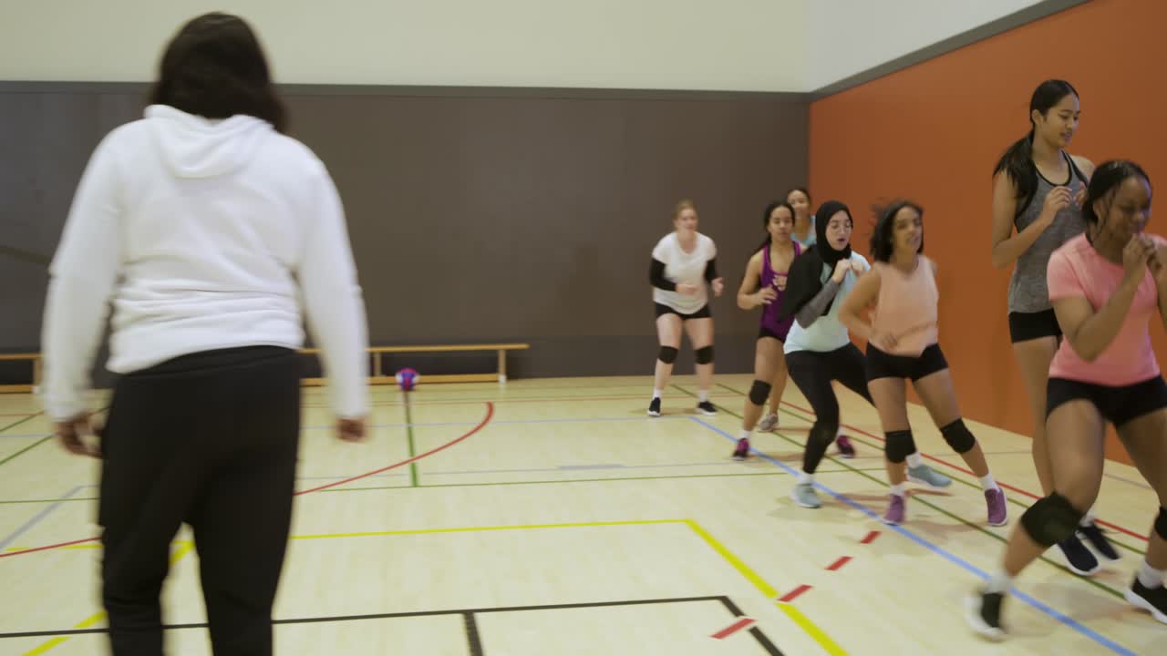 Squadra femminile di pallavolo che esegue saltelli a ginocchia alte per il riscaldamento sul campo da palestra, con l'allenatore che fa segnali, vicino alla rete.