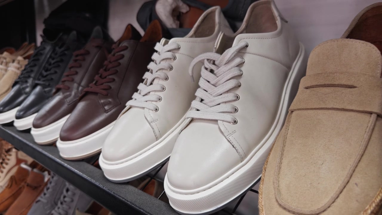 Filmisk 4K-optagelse af herresko pænt arrangeret på metalhylder i en detailbutik. Loafers, oxfords, sneakers og mokkasiner i forskellige farver og materialer udstillet.