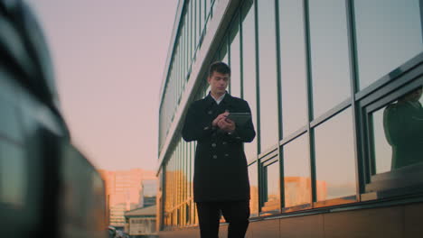 business man in black coat walking close to glass building, focusing back on tablet reflecting on glass, background features blurred building and parked cars