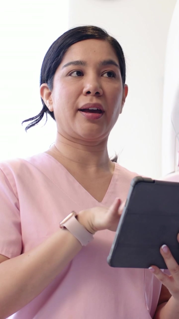 Video verticale: Personale in camice rosa che guarda in basso, confermando le informazioni sul tablet in clinica accanto alla finestra.