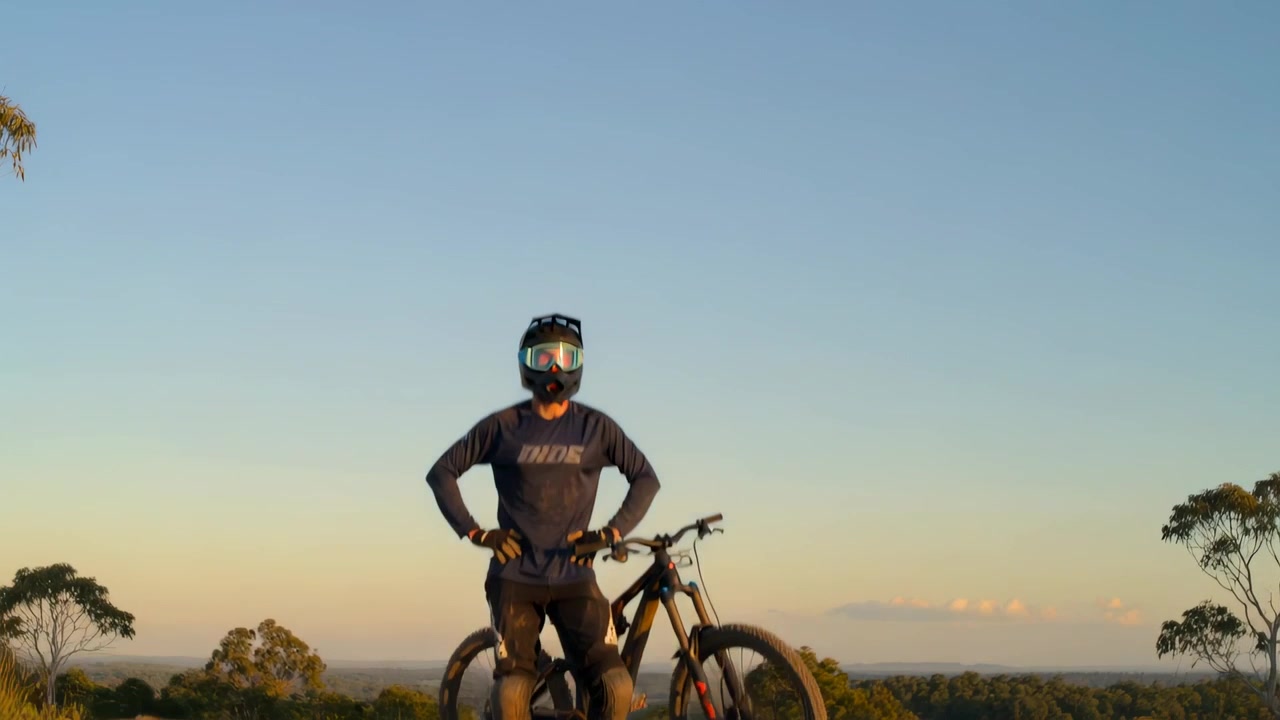 Telecamera che si allontana rivelando un motociclista con casco e moto su una cresta al tramonto, mostrando una valle distante.