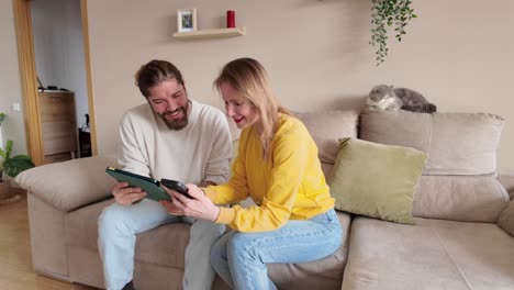 couple laughing while looking at a tablet with a cat nearby