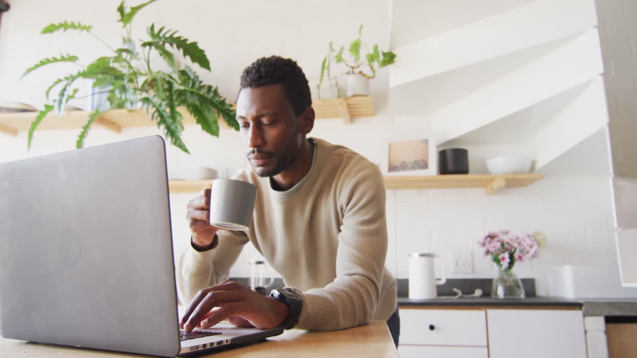 Happy african american man leaning on countertop in kitchen, using laptop and drinking coffee