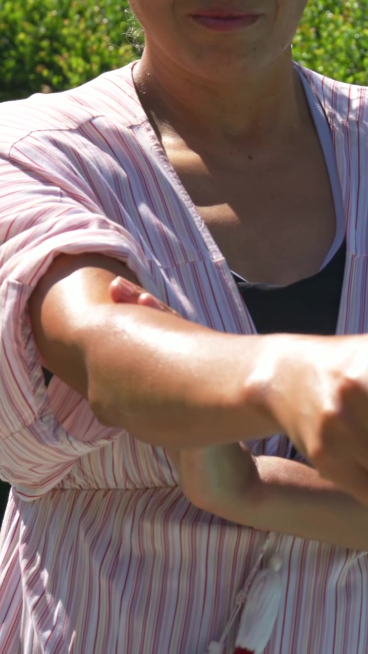 Video vertical: Levantando el brazo derecho, mujer frotándose el antebrazo en el jardín, arreglándose el vestido de rayas y las gafas de sol