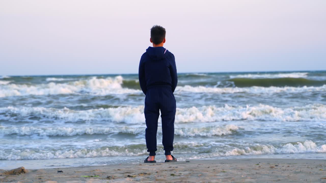 Teenage boy looking at the sea. Backside view of a child in clothes standing on a sandy beach near the sea and looking on waves in summer evening.