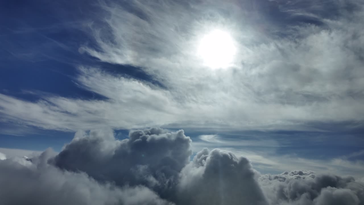 An immersive aerial cloudscape through the pilot’s eyes from a jet cockpit, flying over cottony clouds with a shinig sun veiled by ethereal cirrus clouds.