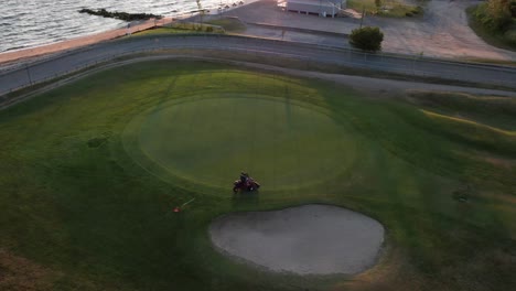 Eine-Luftaufnahme-Einer-Person,-Die-Auf-Einem-Rasenmäher-Fährt-Und-Bei-Sonnenaufgang-Das-Gras-Eines-Golfplatzes-Neben-Einem-Strand-Schneidet