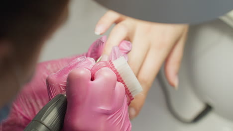 close up top view of nail technician wearing pink gloves and protective mask using brush and electric file to carefully clean and shape customer's fingernail during precise manicure procedure
