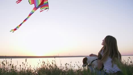 young happy woman and het little dog sitting with flying kite on a glade at sunset