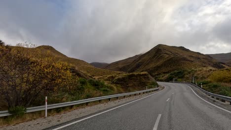 a vehicle travels a curving rural road through hilly farmland, surrounded by golden grass and mountains under overcast evening skies, captured in smooth forward motion