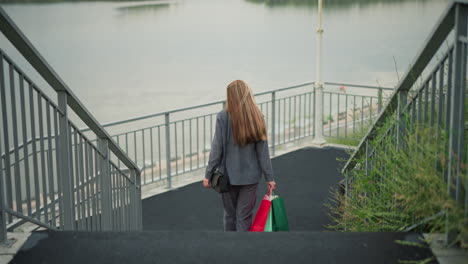 back view of lady carrying colorful shopping bags walking down stairs while adjusting her hair, turning slightly left near bottom of stairs, background features a calm river, lamp post, and greenery