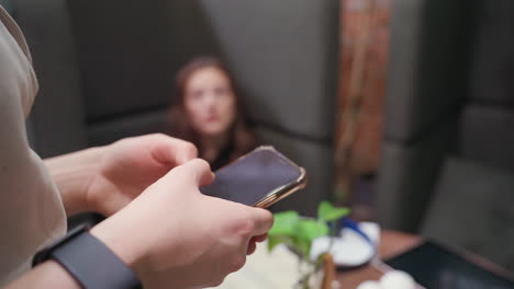 woman stands in foreground holding phone with two hands, wearing wristband, while another woman is seen out of focus in background, sitting in booth area with soft lighting and relaxed atmosphere