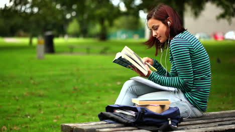 Pretty-student-sitting-on-bench-reading-a-book-listening-to-music