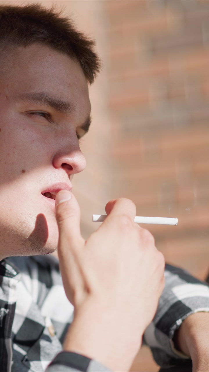 Primer plano de un hombre joven con cabello castaño corto, vistiendo una camisa a cuadros blanca y negra, sosteniendo un cigarrillo electrónico. El fondo presenta ladrillos borrosos, indicando un entorno urbano al aire libre.