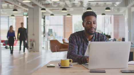 Hombre-De-Negocios-Trabajando-En-Una-Computadora-Portátil-En-El-Escritorio-Con-Una-Bebida-Caliente-En-Una-Oficina-Moderna-Con-Colegas-En-Segundo-Plano