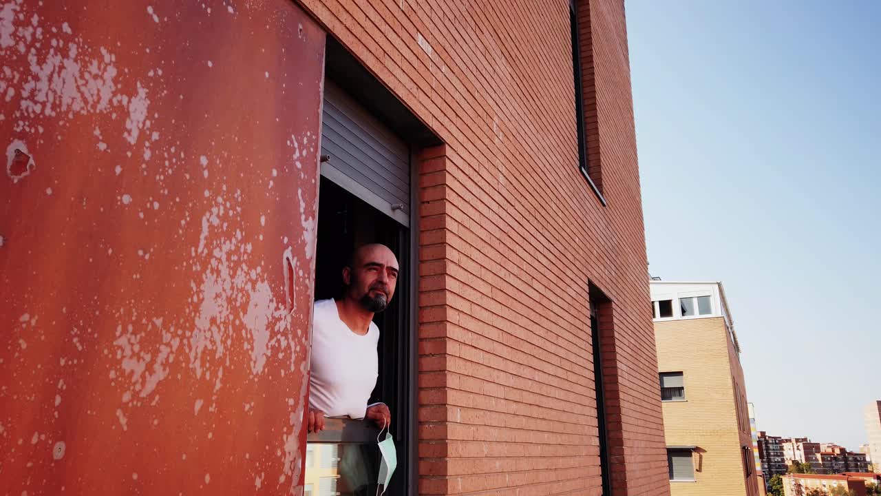 A shot of a man peeking out a window wearing a disposable face mask, taking it off and taking deep breath