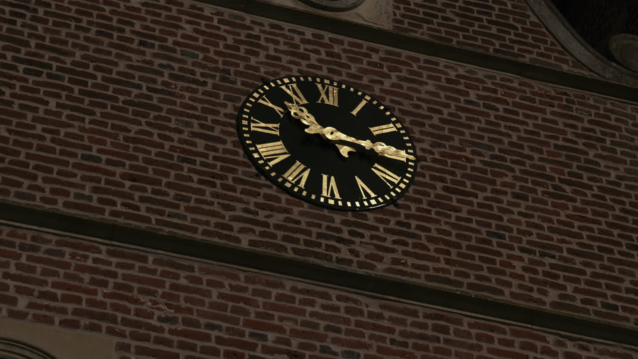 A detailed view of a church clock with golden Roman numerals and hands on a black face, set against the brick walls of a historic building, highlighting timeless European architecture