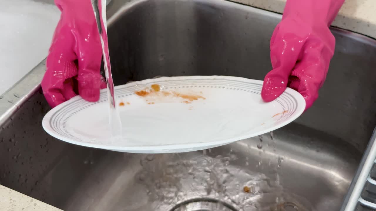 Hands in pink gloves rinse a dirty plate under running water in a stainless steel kitchen sink, removing food residue with gentle scrubbing