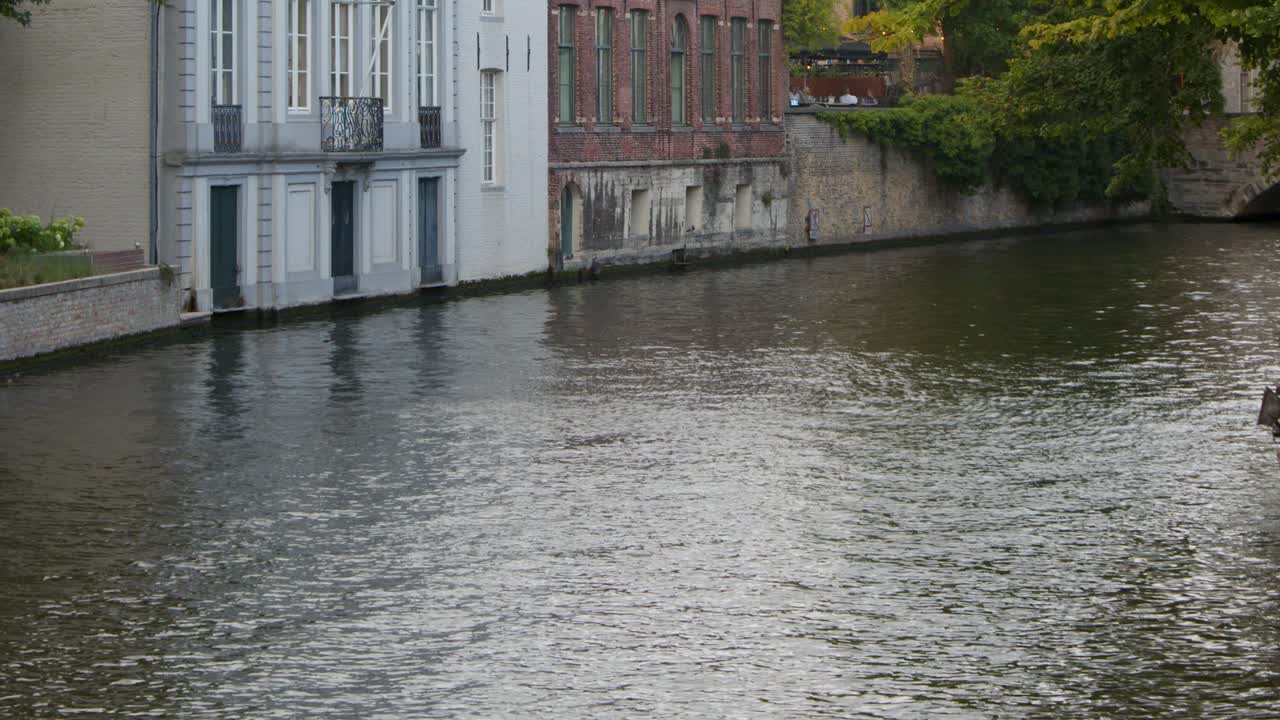 Camera slowly pans along tranquil Bruges canal, highlighting historic architecture, autumn trees, soft daylight