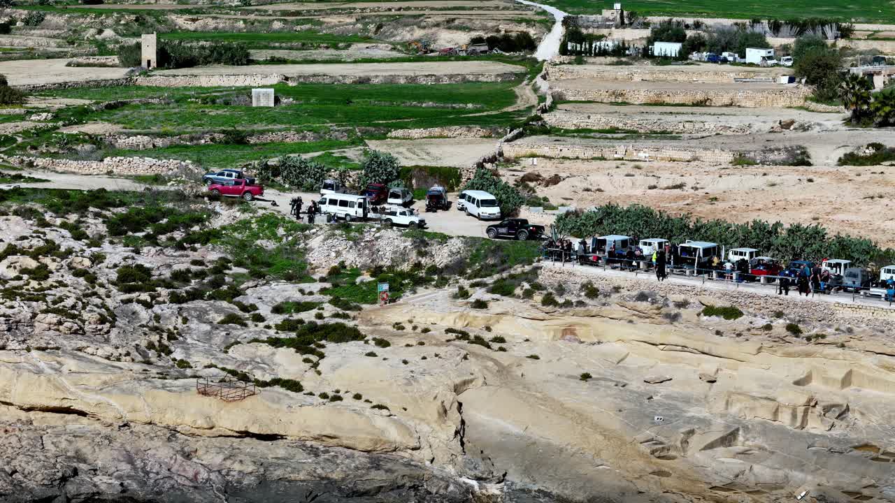 Ripresa panoramica dall'alto di un'area di parcheggio rustica immersa tra i campi verdi terrazzati e le scogliere calcaree dorate di Gozo, Malta, sotto un luminoso sole primaverile con cieli azzurri e sereni e tonalità terrose.