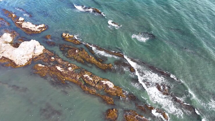 Drone footage captures turquoise waves breaking over rugged rocks along the St Andrews coastline, with bright natural daylight and a gentle camera pan