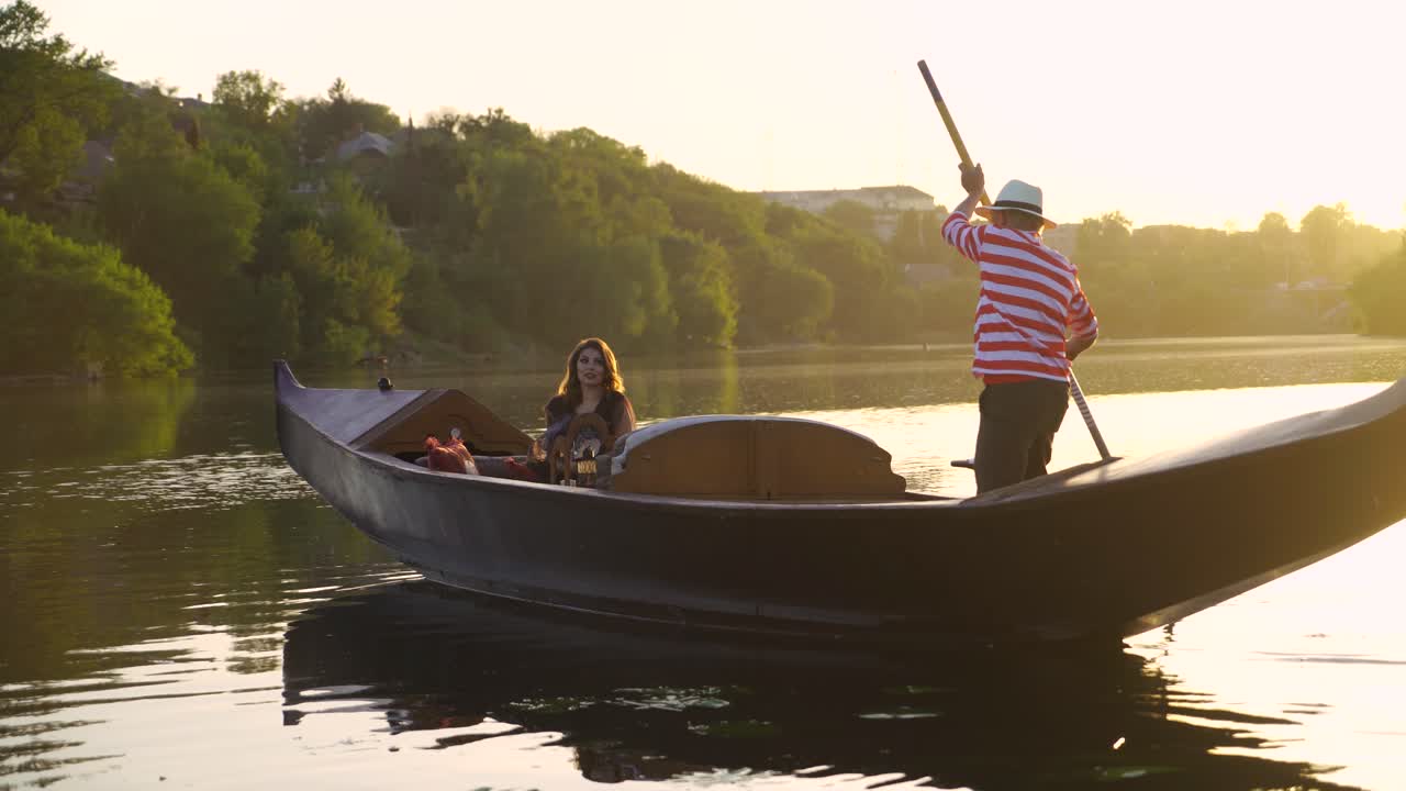 Beautiful woman sitting in a boat and a man rowing by oar on the natural background at sunset.Traditional venetian rowing boat with the gondolier and a female in the evening river.