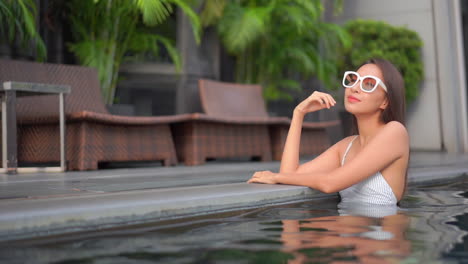 pequeña mujer asiática en traje de baño y gafas de sol de pie en el agua de la piscina y mirando alrededor, fotograma completo