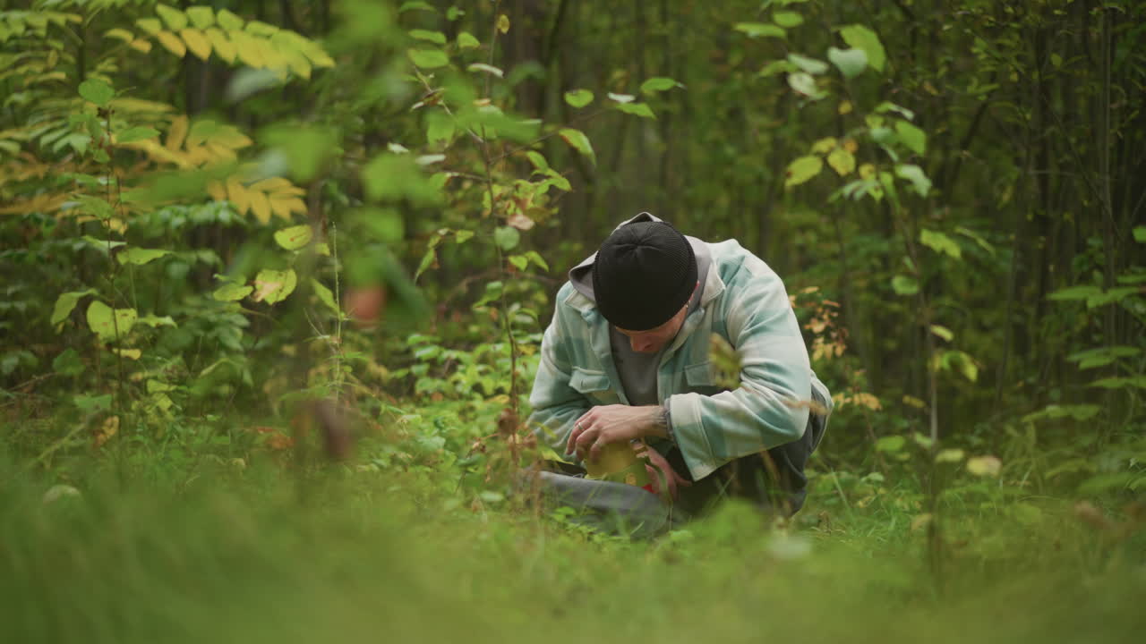 Kaukasisk turgåer huker seg ned i tett skog og inspiserer kompass og ryggsekk, mosegrodd bakke og grønt løvverk, lue og jakke, fokusert sanker som plukker sopp, utendørs guide reparerer reim