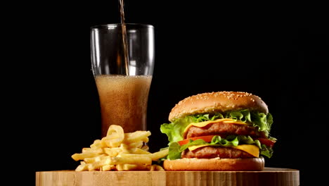 berger and french fries lie on a wooden board on a black background, close-up poured soda in a glass