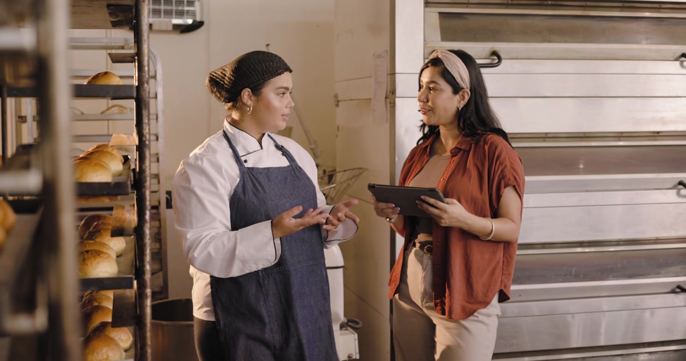 Squadra femminile di panettieri alle rastrelliere, con la panettiera in giacca da chef che indica mentre la manager usa il tablet per controllare la produzione.