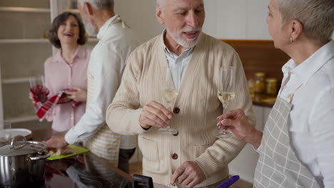 older man and woman friends toasting with a glass of wine, while a couple of older friends cooking in the kitchen in the background