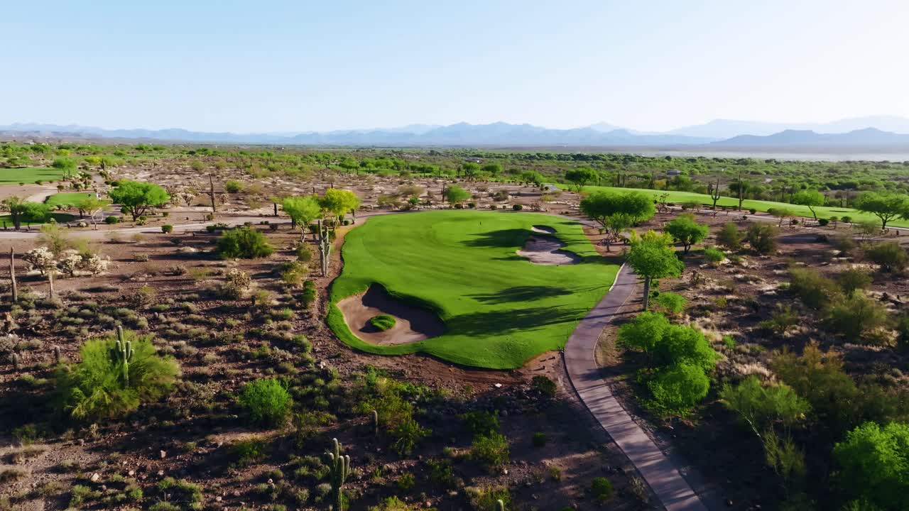 Aerial push-in shot of a desert golf green with surrounding bunkers and cart path, framed by distant mountains