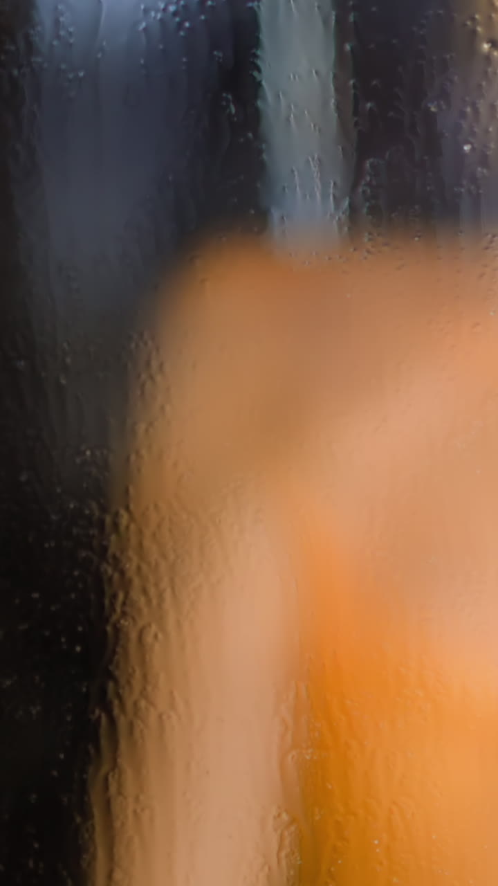 Young woman takes shower washing body behind water drops flowing down transparent glass cabin door in bathroom at home slow motion closeup