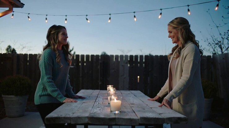 Arranging two women placing glass candles on wood table at dusk, creating ambiance in teal sweater