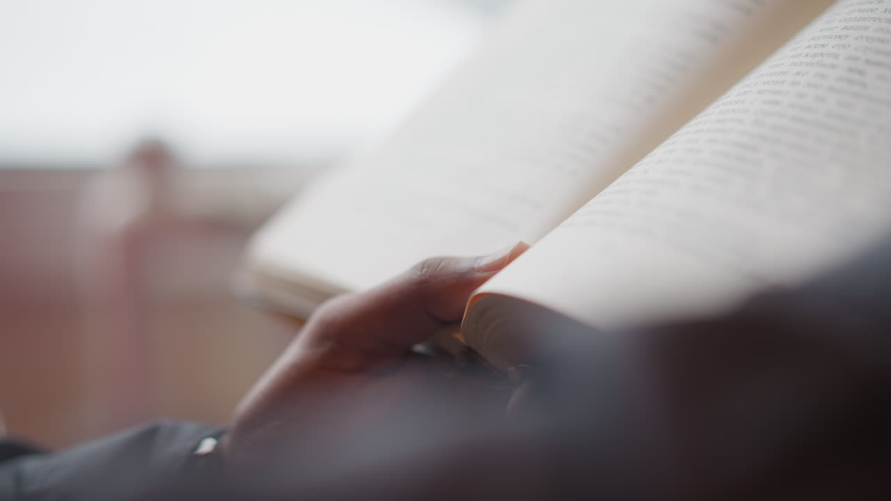 Close-up of male hand gently turning page of open book in soft natural light, text in focus, background blurred, capturing quiet moment of reading, study, or thoughtful engagement